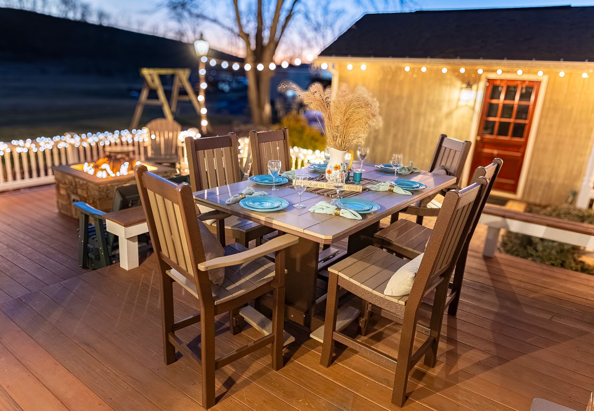 Outdoor dining set on a wooden deck with lit string lights, shed, and fire pit at dusk.