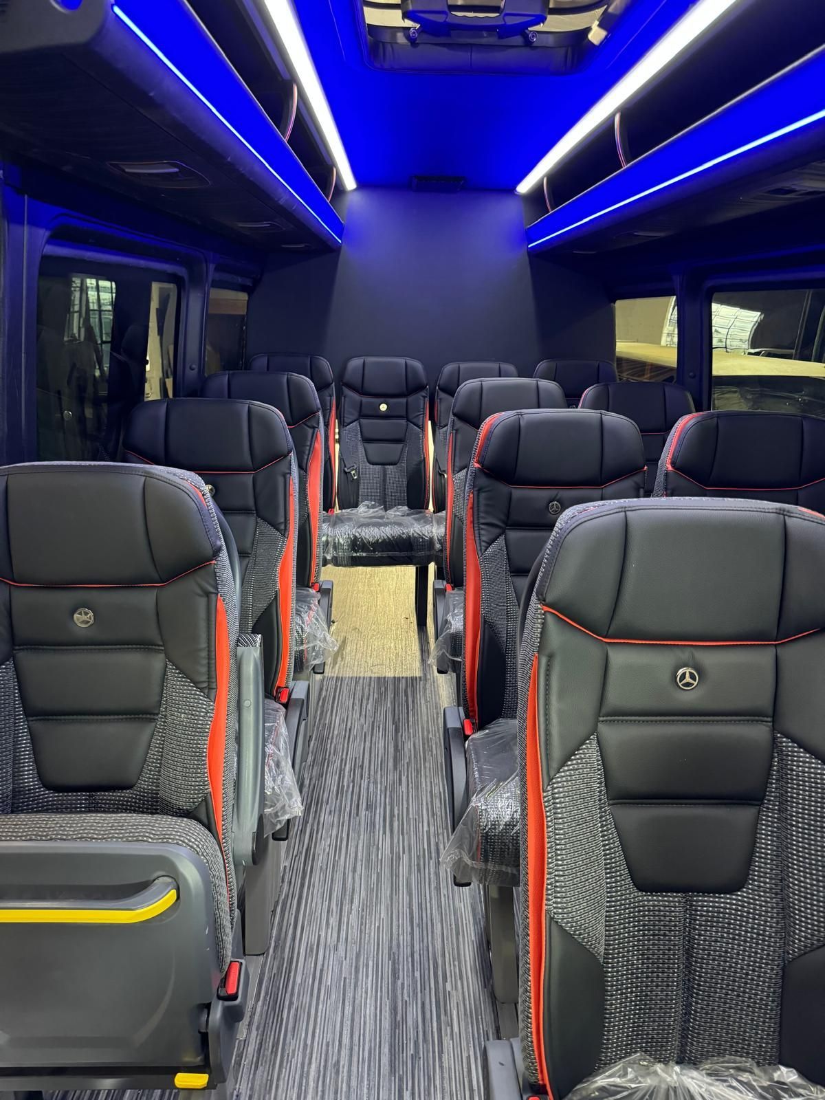 Interior of a dark bus with rows of black seats and blue overhead lighting.