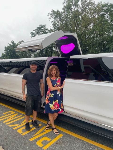 Two people stand by a white limousine with an open door. The woman wears a floral dress, man wears a t-shirt and shorts.