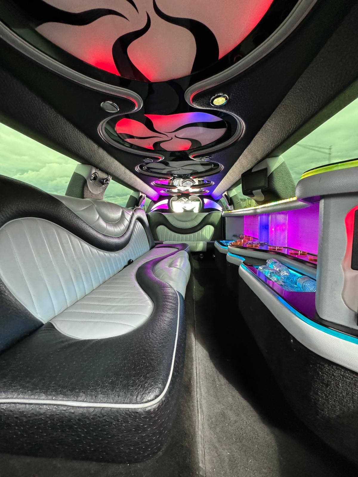 Inside a limousine with black, gray, and white seating, bar with purple lights, and decorative ceiling lights.