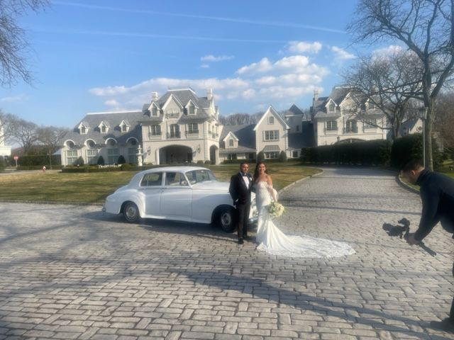 Wedding couple poses by white car in front of large mansion; photographer films.