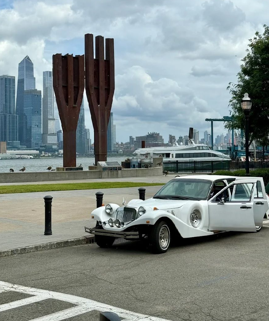 White vintage limo parked near a waterfront with sculptures and a city skyline.
