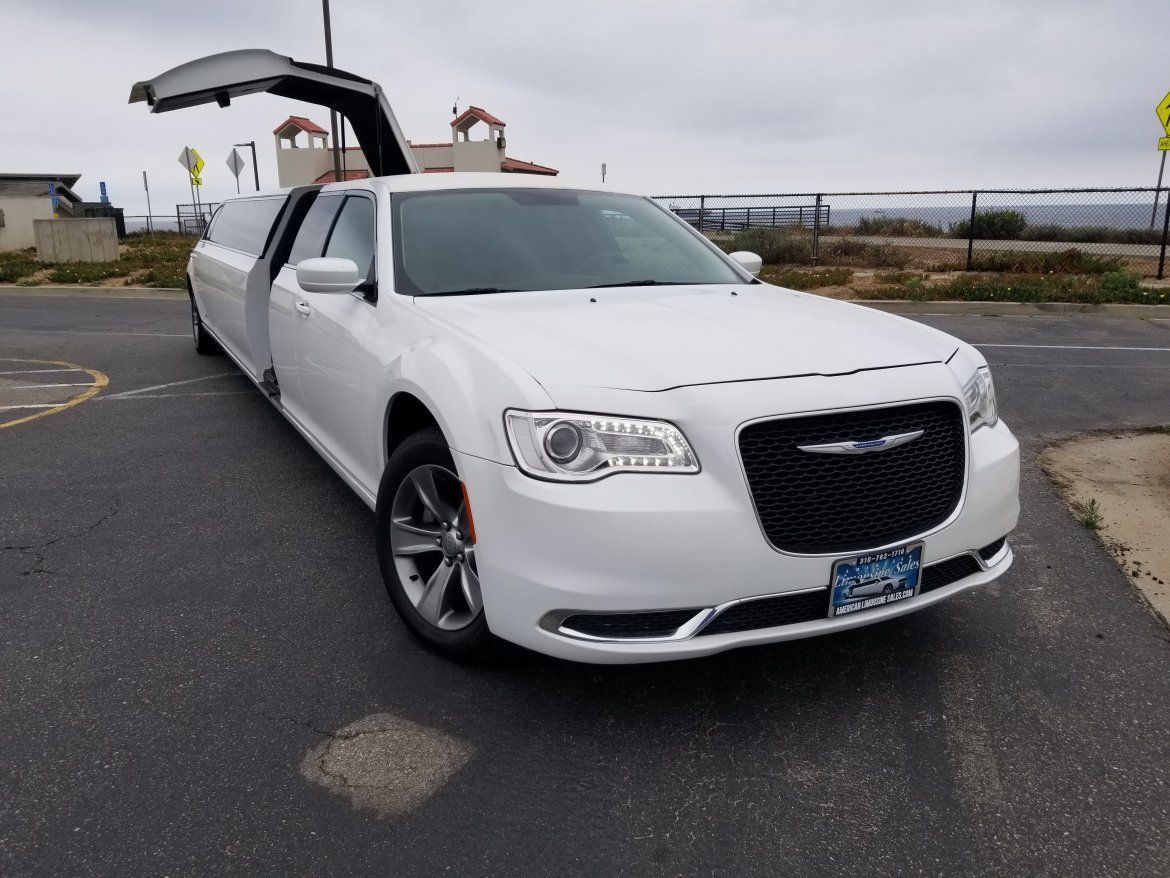 White Chrysler 300 limousine with gullwing doors open on a road. Cloudy sky in the background.
