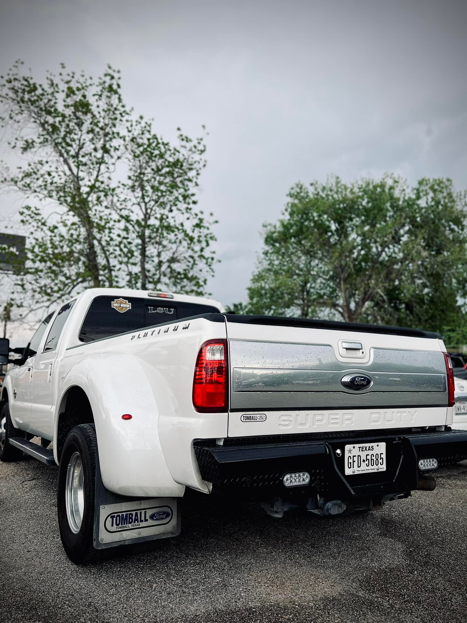 White Ford dually truck, parked on gravel, rear view. Silver tailgate, black bumper, mud flaps.