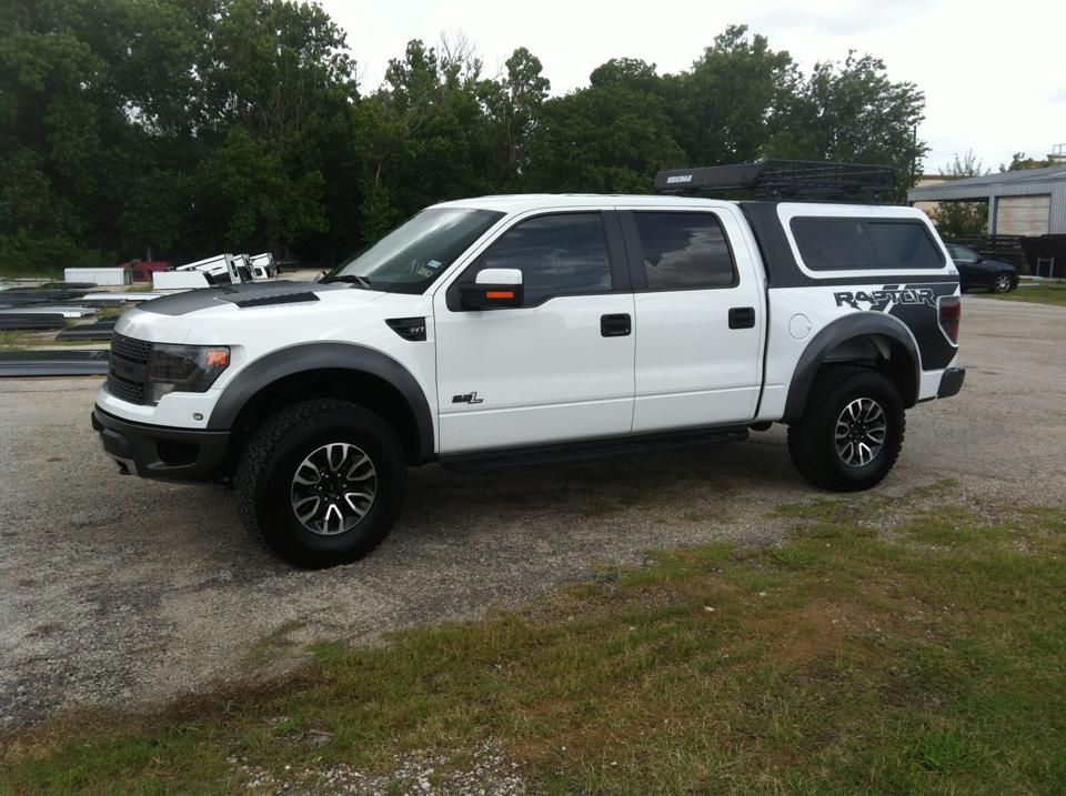 White Ford Raptor truck with black accents, black wheels, and a camper shell parked outdoors.
