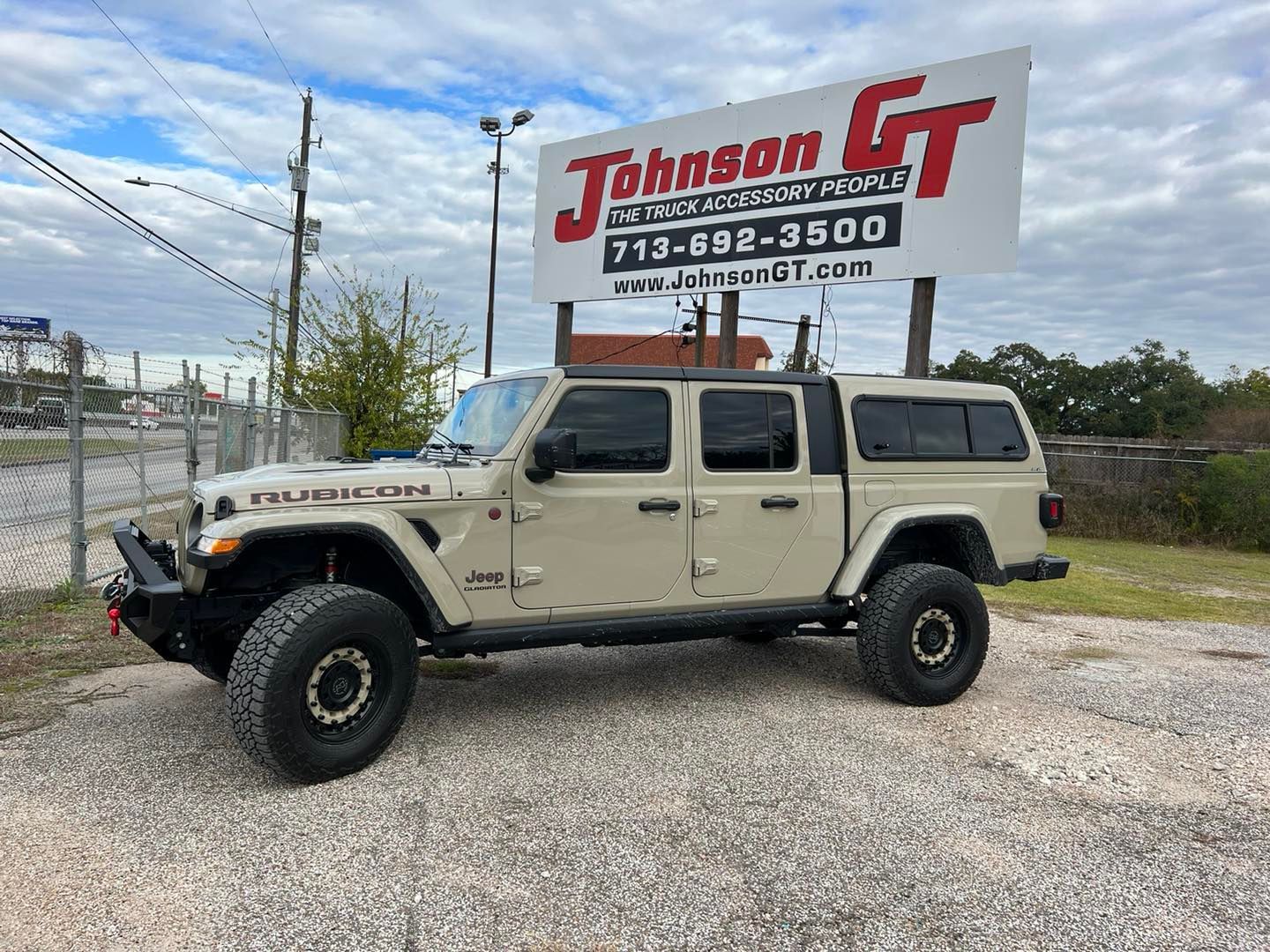 Tan Jeep Gladiator truck with black accessories and a camper shell, parked in a lot in front of a dealership sign.