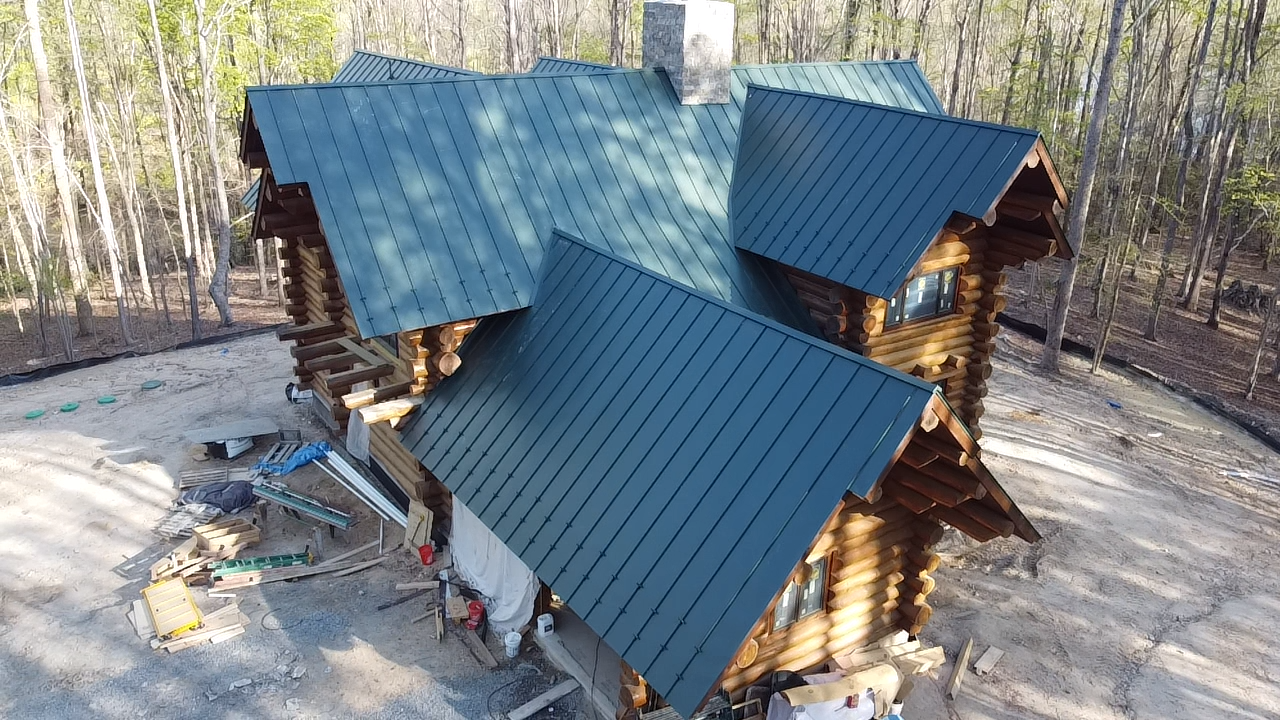 An aerial view of a log cabin with a green roof in the middle of a forest.