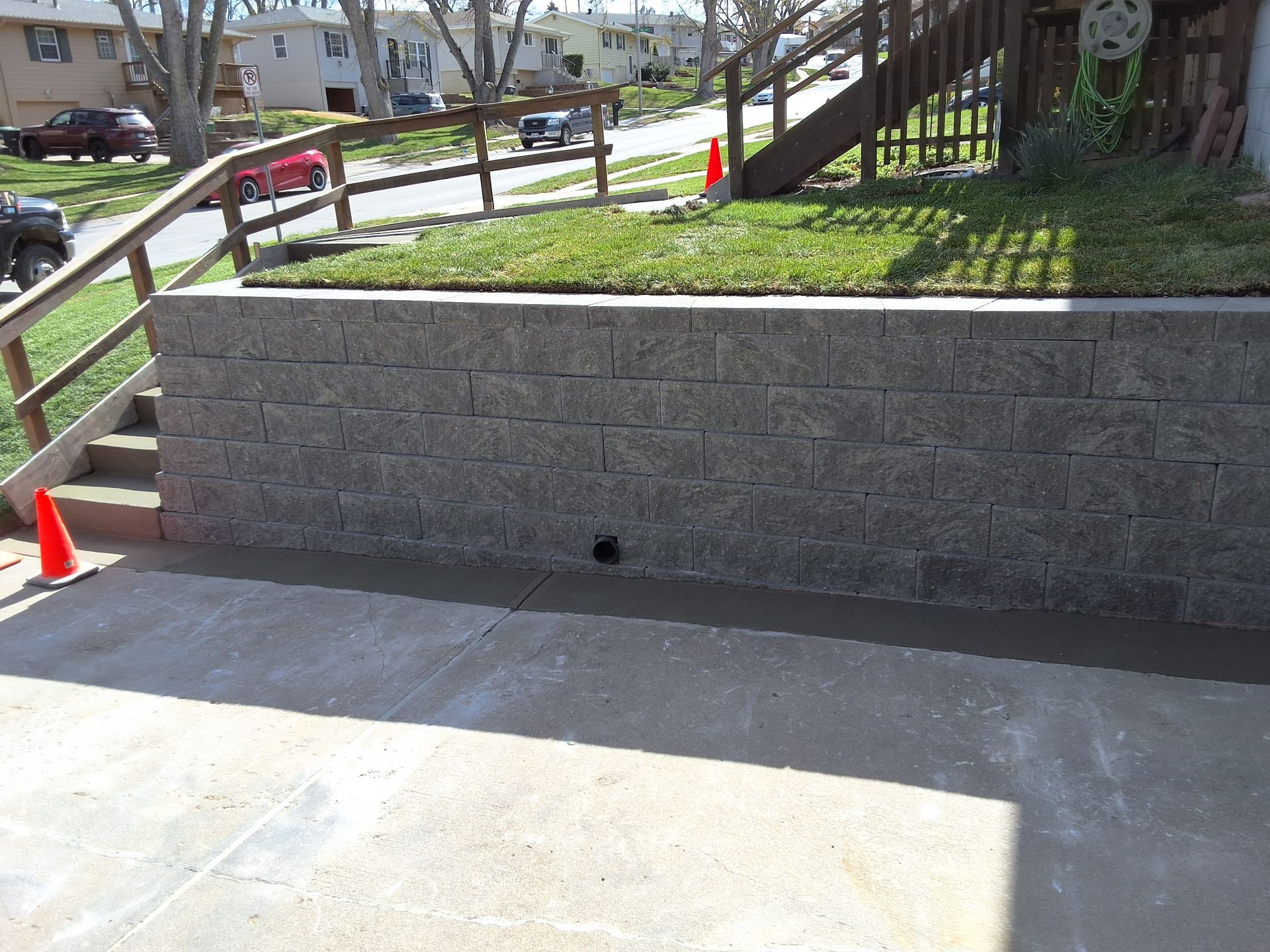 A newly constructed gray concrete block retaining wall next to a concrete driveway, wooden stairs, and a grassy yard.