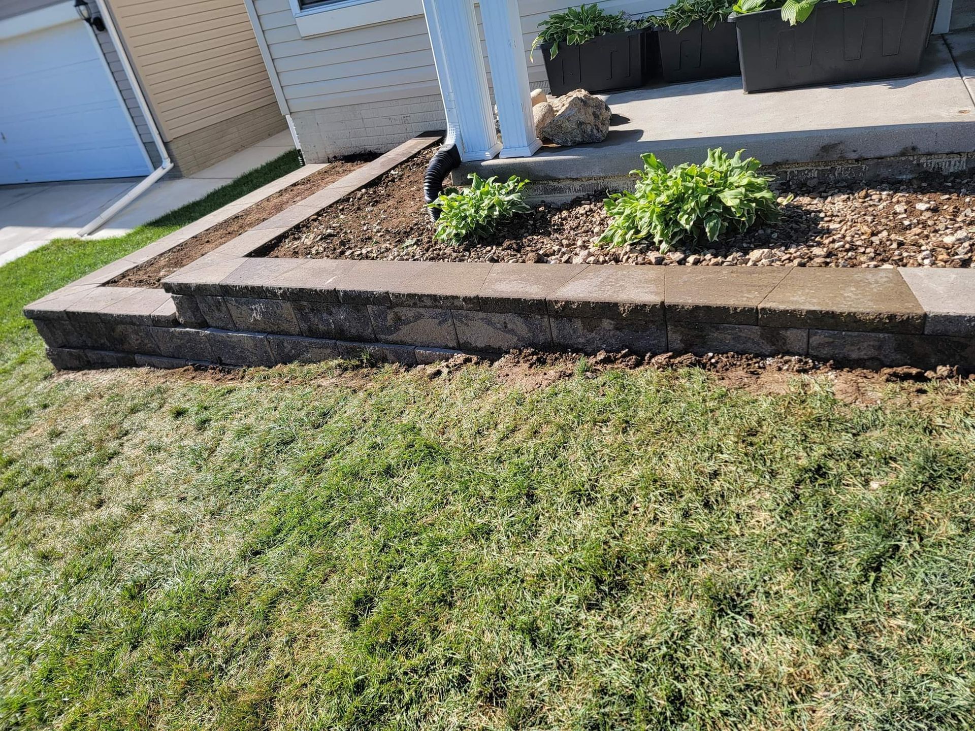 A gray stone retaining wall borders a mulch garden bed with small green plants in front of a house.