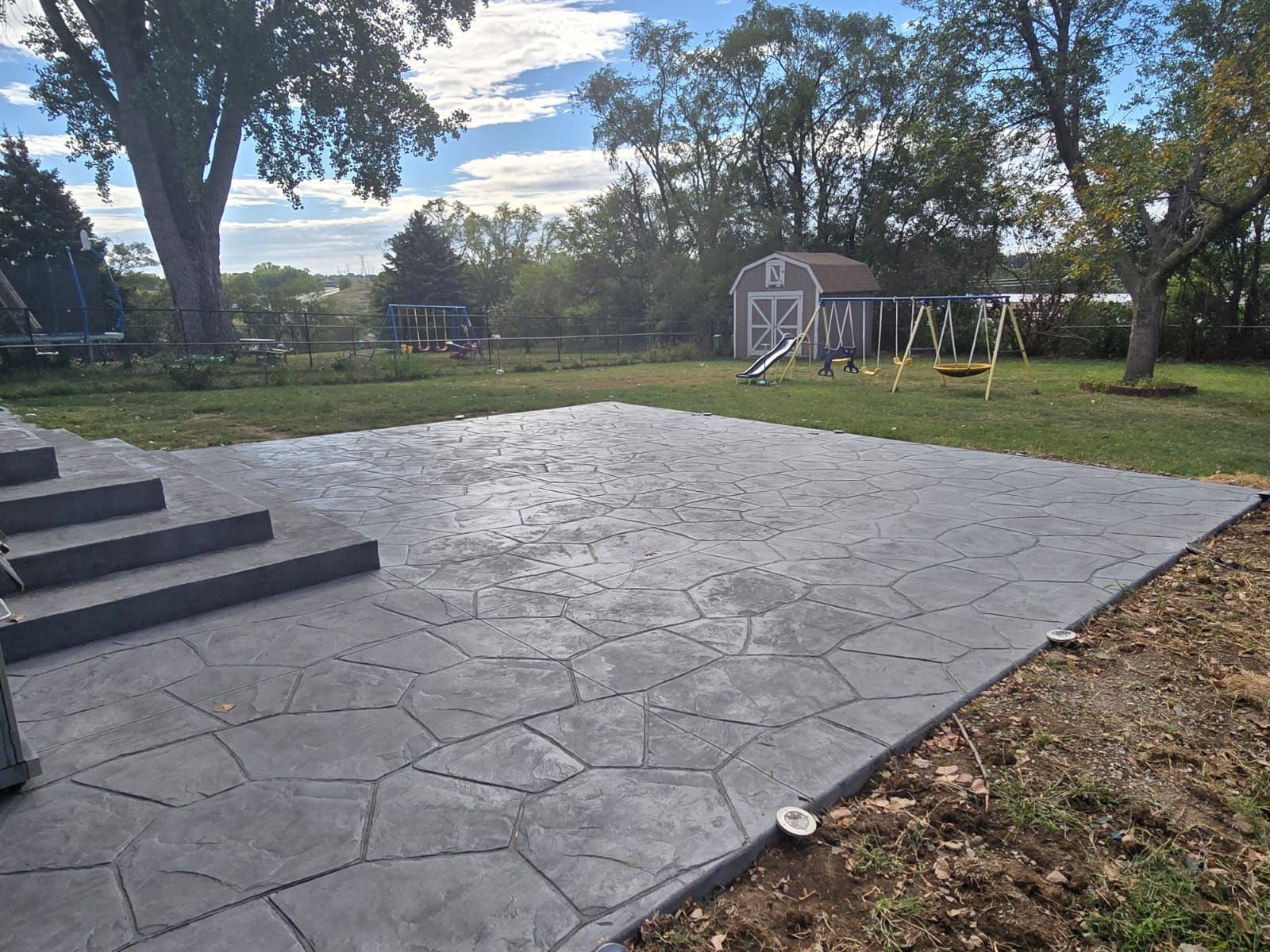 A grey, stamped concrete patio with stone-patterned tiles sits beside stone steps in a grassy backyard with a shed.