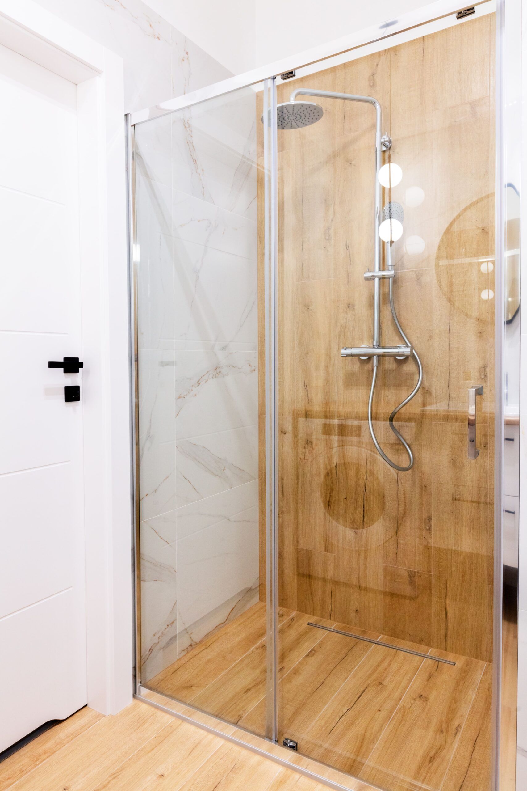 Shower with wooden-look tiled walls and floor, glass enclosure, chrome fixtures, and a white door.