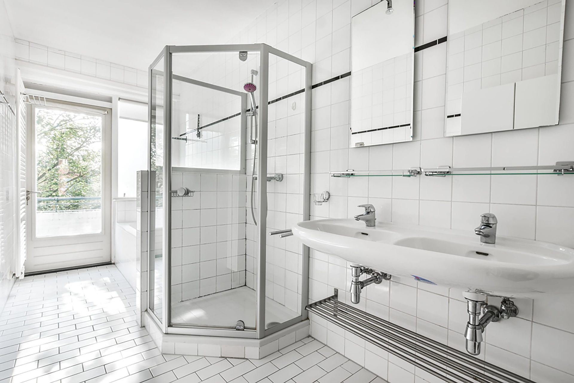 Bright white bathroom with glass shower, double sink, and a door to the outdoors.