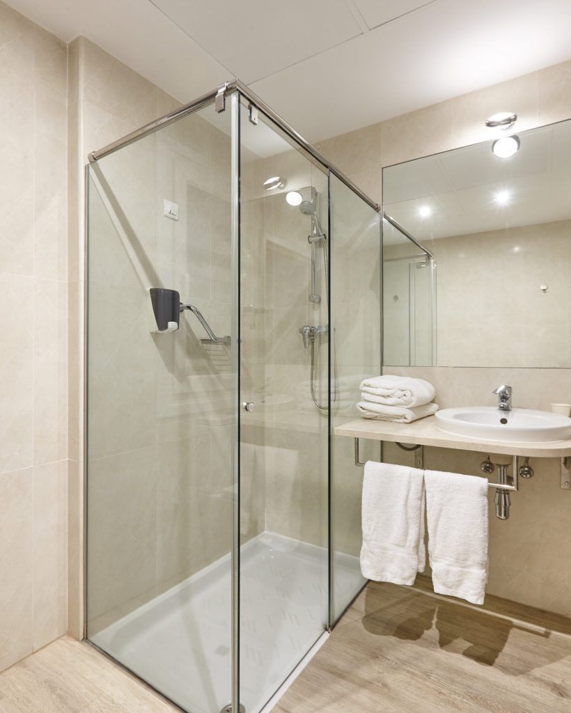Corner shower with clear glass doors in a bathroom. White sink, towels, and neutral-toned walls.