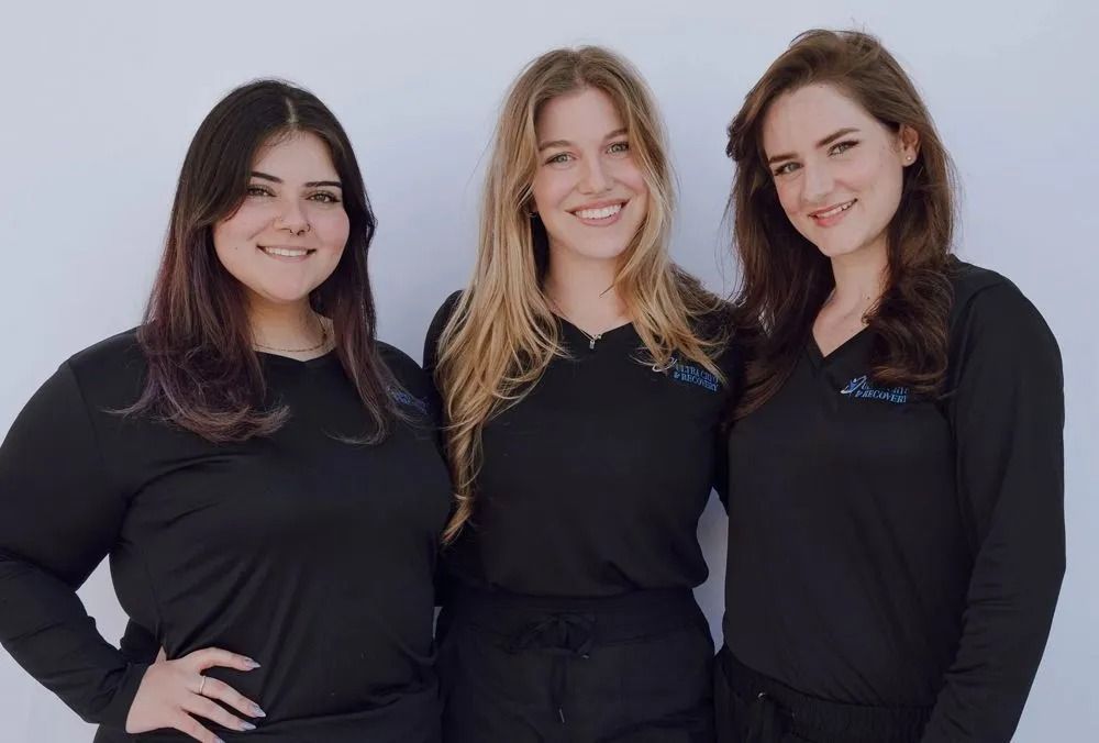 Three women in black shirts smiling in front of a white wall.