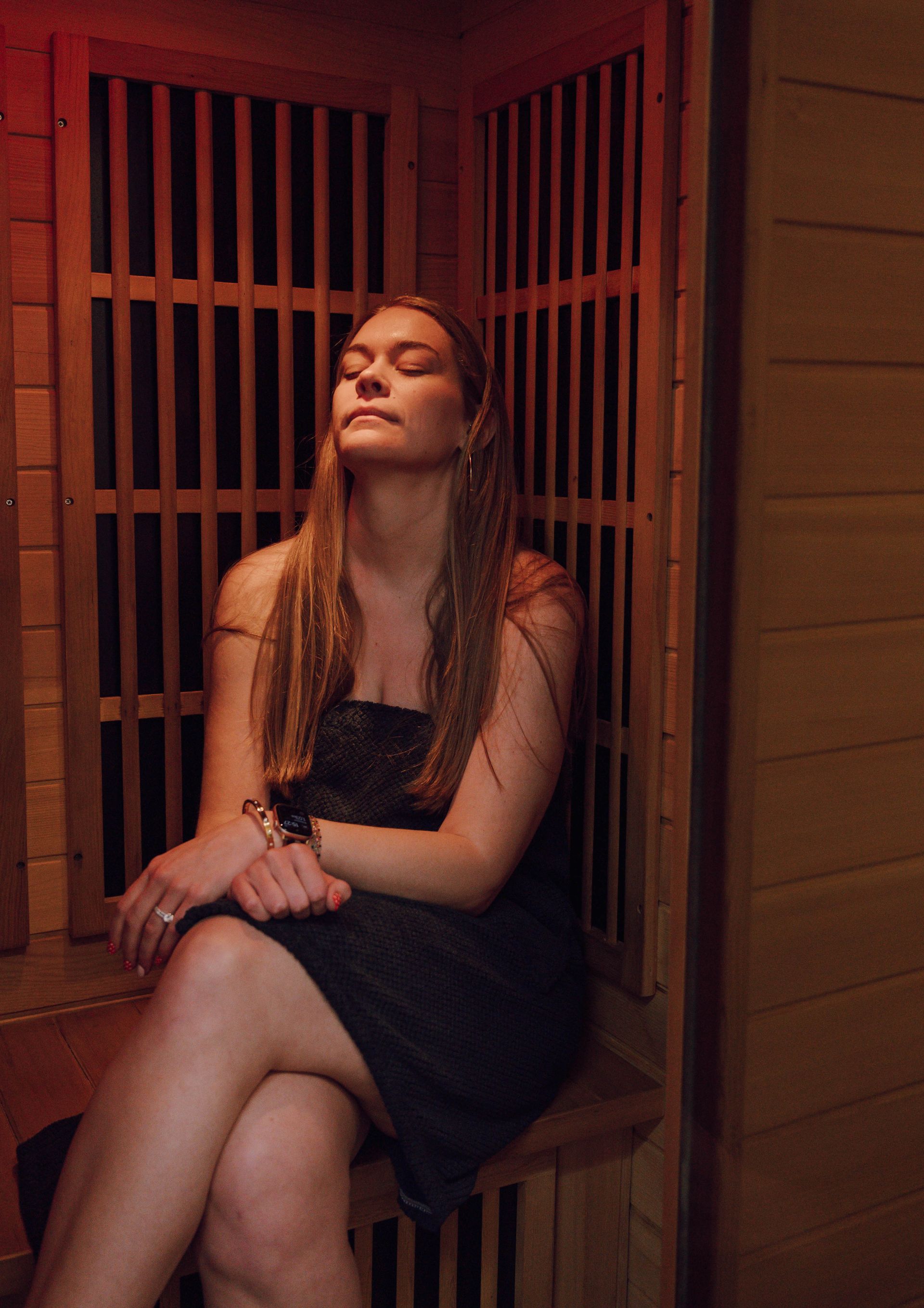 A person sits with eyes closed, relaxing inside a wooden sauna with warm ambient lighting.