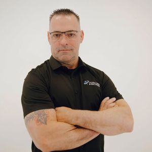 Man in black shirt with arms crossed, wearing glasses. Tattoo on left arm.
