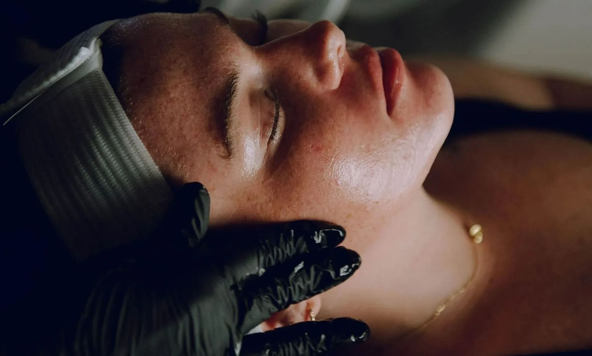 Person receiving a facial treatment with black gloved hands on their face, lit in dim light.