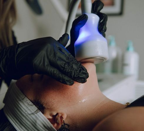 Person receiving a facial treatment with a glowing blue light device; a gloved hand applies the device to the neck.