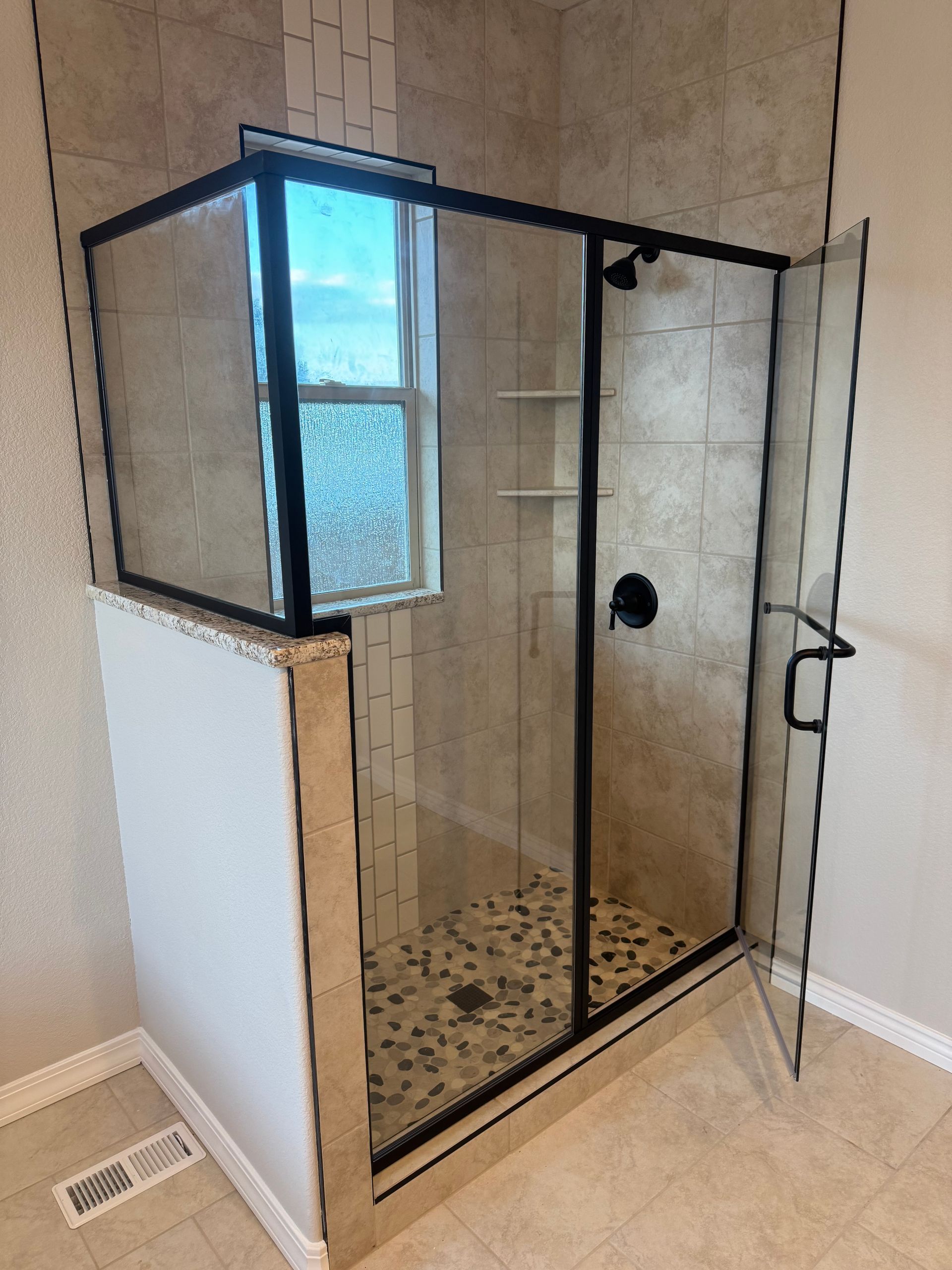 Modern shower with glass doors, black frame, pebble floor, and small window.