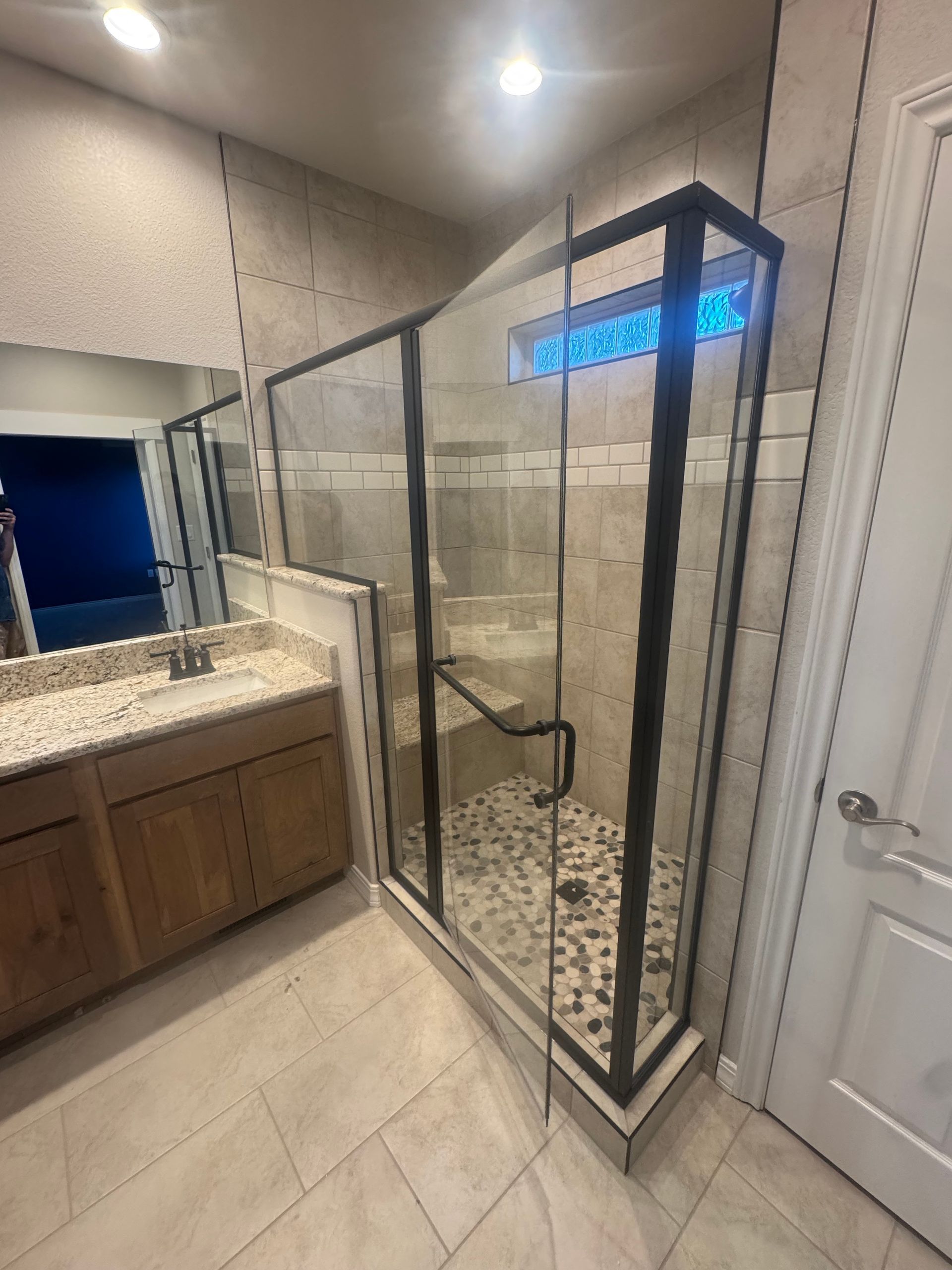 Bathroom with glass-enclosed shower, black framing, pebble floor, beige tile walls. 