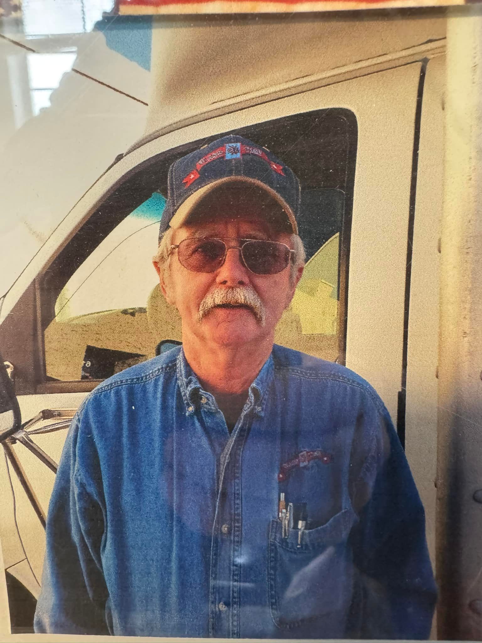 Man in blue denim shirt and cap, standing in front of a white van. He has a mustache and glasses.