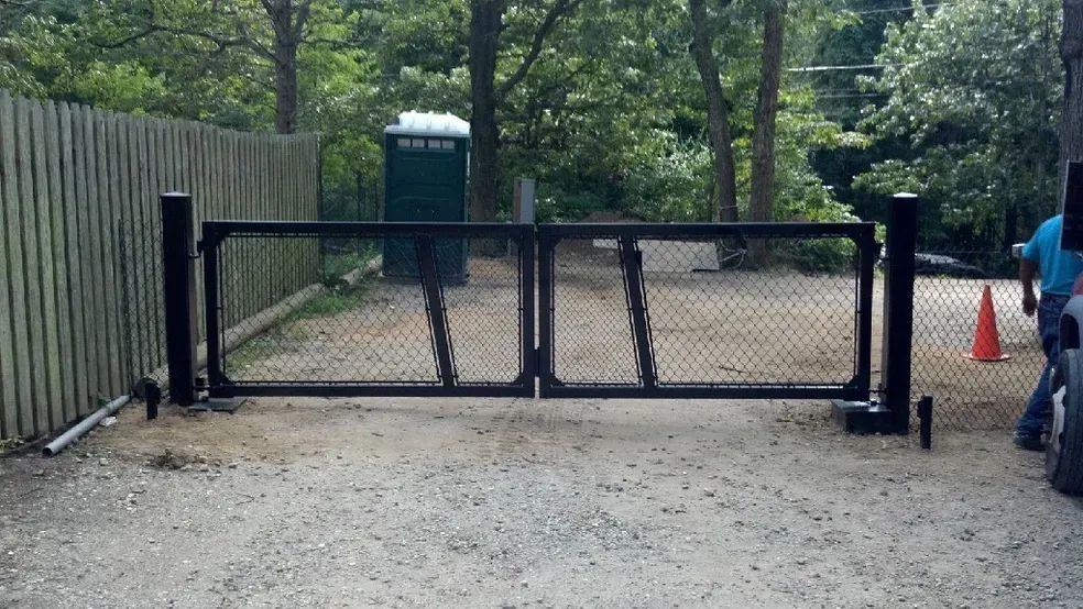 A man is standing in front of a gate in a gravel driveway.