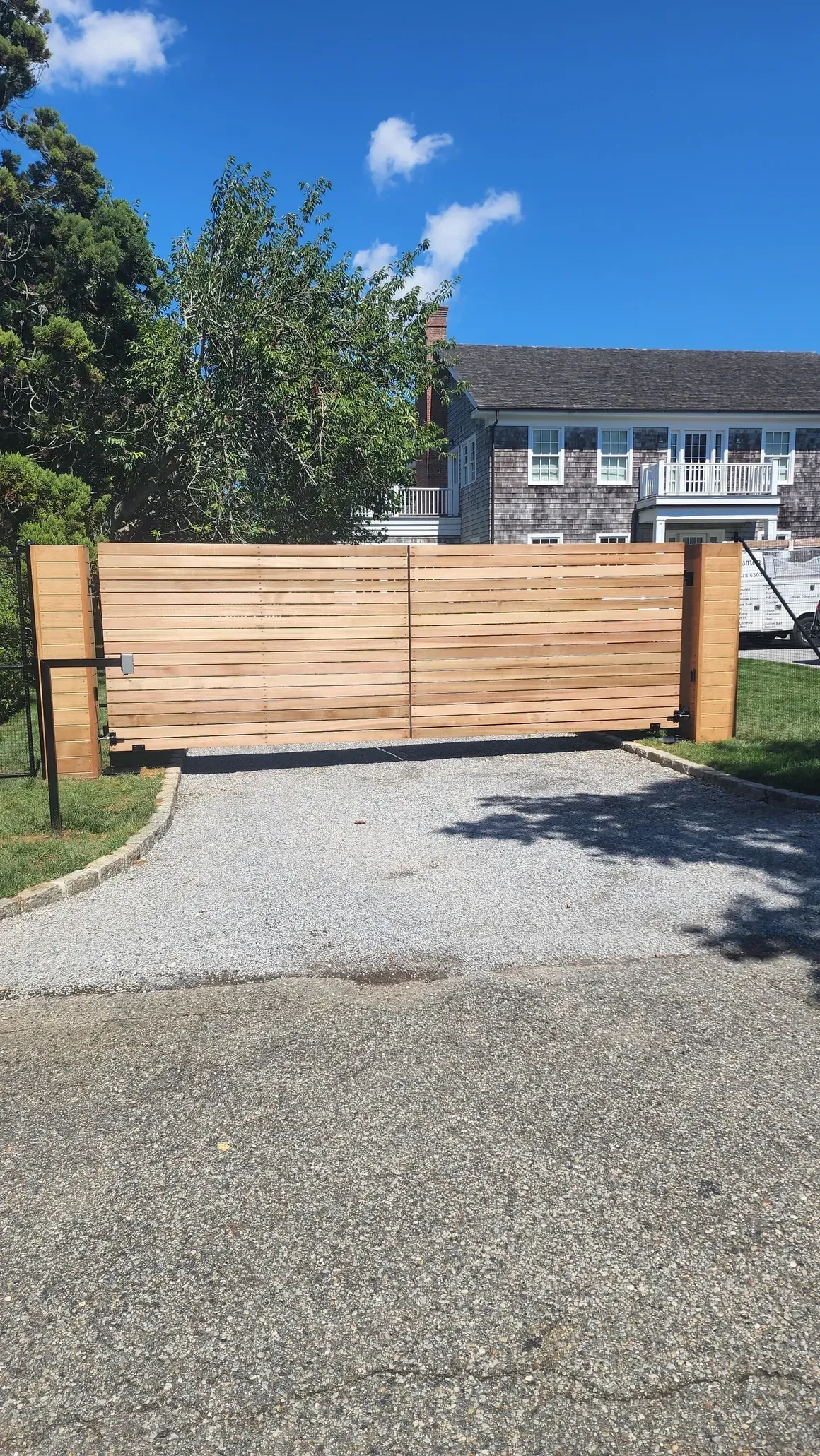 A wooden gate is sitting on the side of a gravel driveway in front of a house.