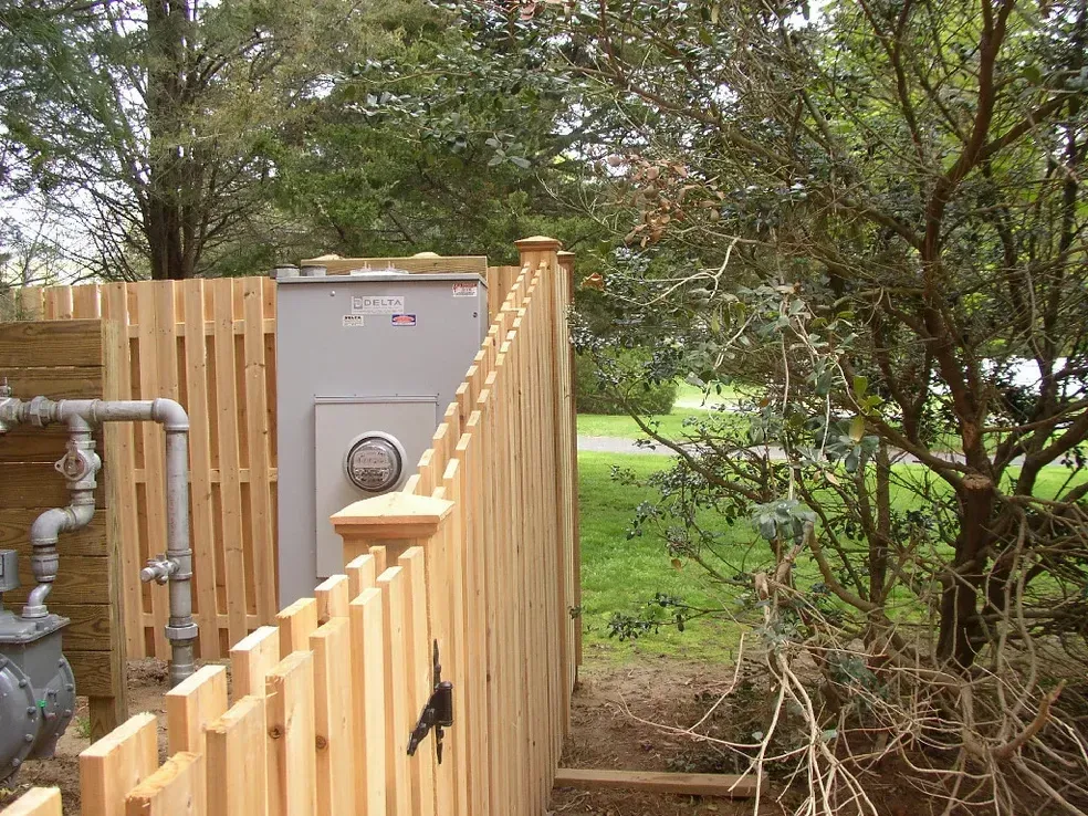 A wooden fence surrounds a utility box in a yard