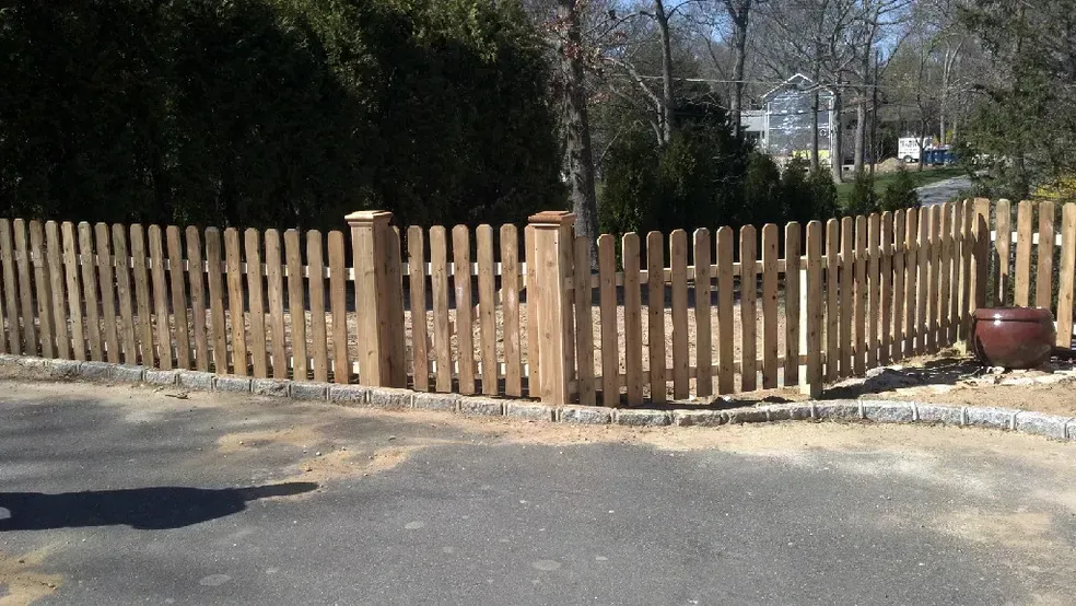 A wooden picket fence surrounds a driveway with trees in the background