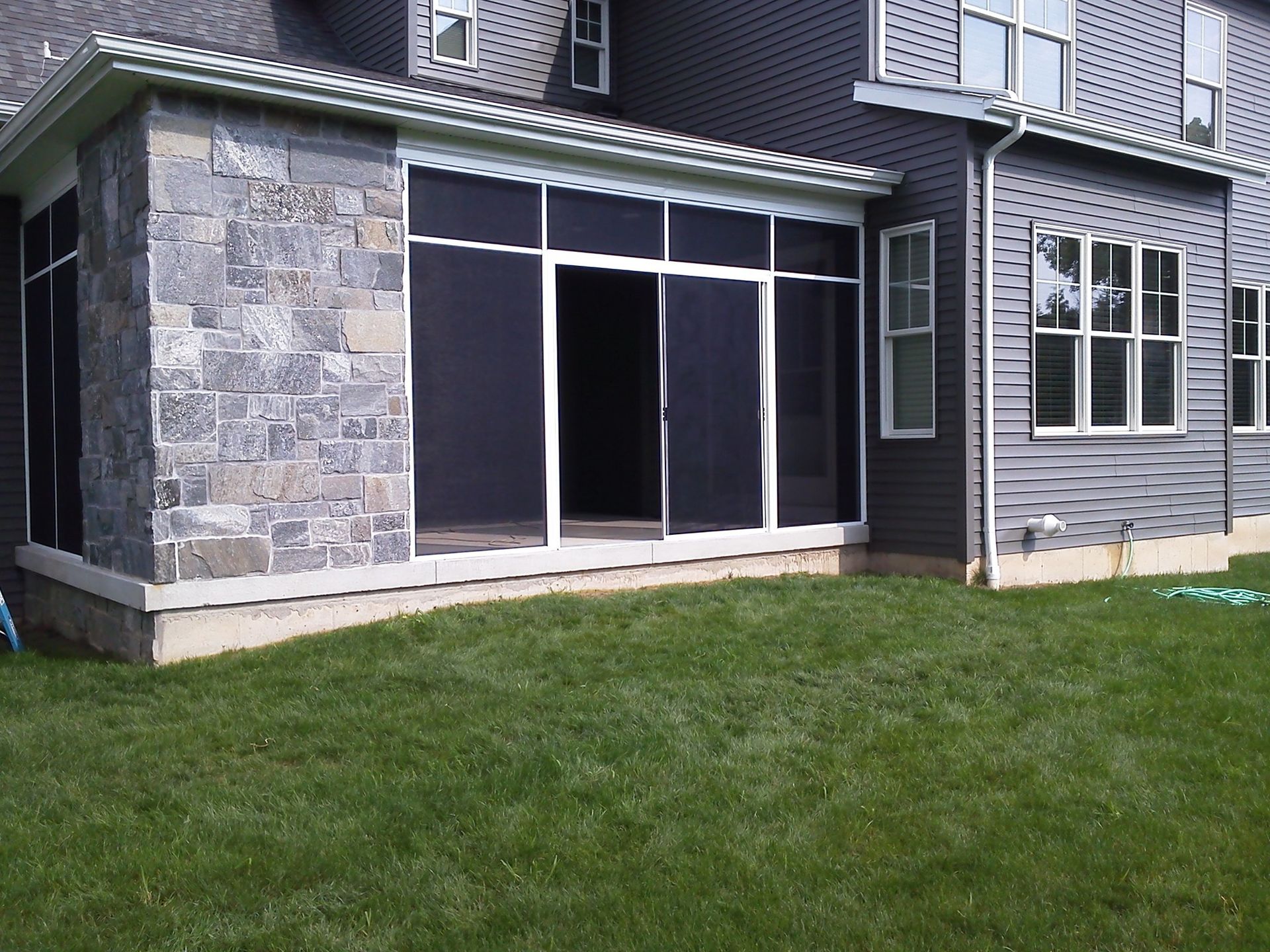 Screened-in porch attached to a house with gray siding and stone accents, overlooking a green lawn.