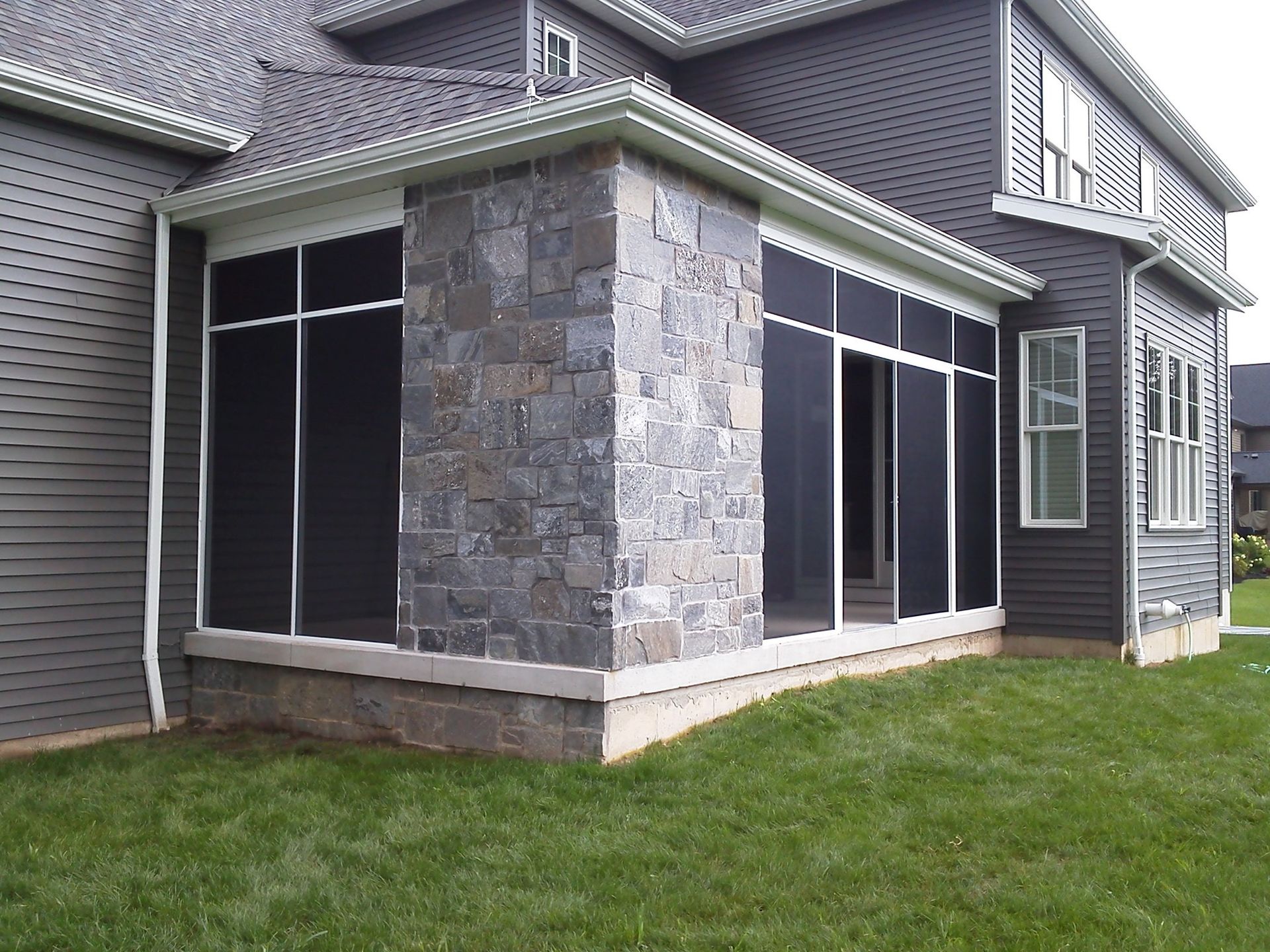 Screened porch with stone wall, attached to a gray house, on a green lawn.
