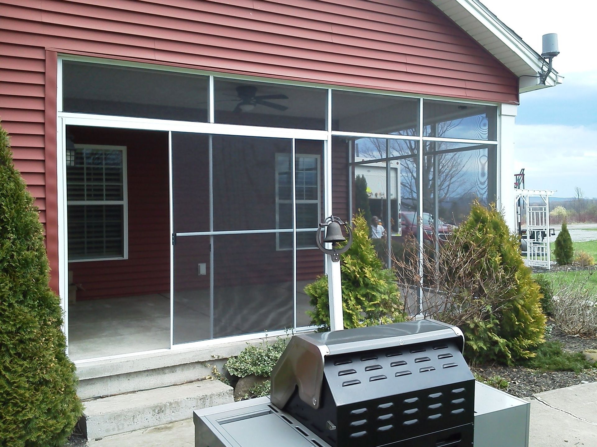 Screened-in porch with a grill, attached to a red house, with some trees and a cloudy sky in the background.