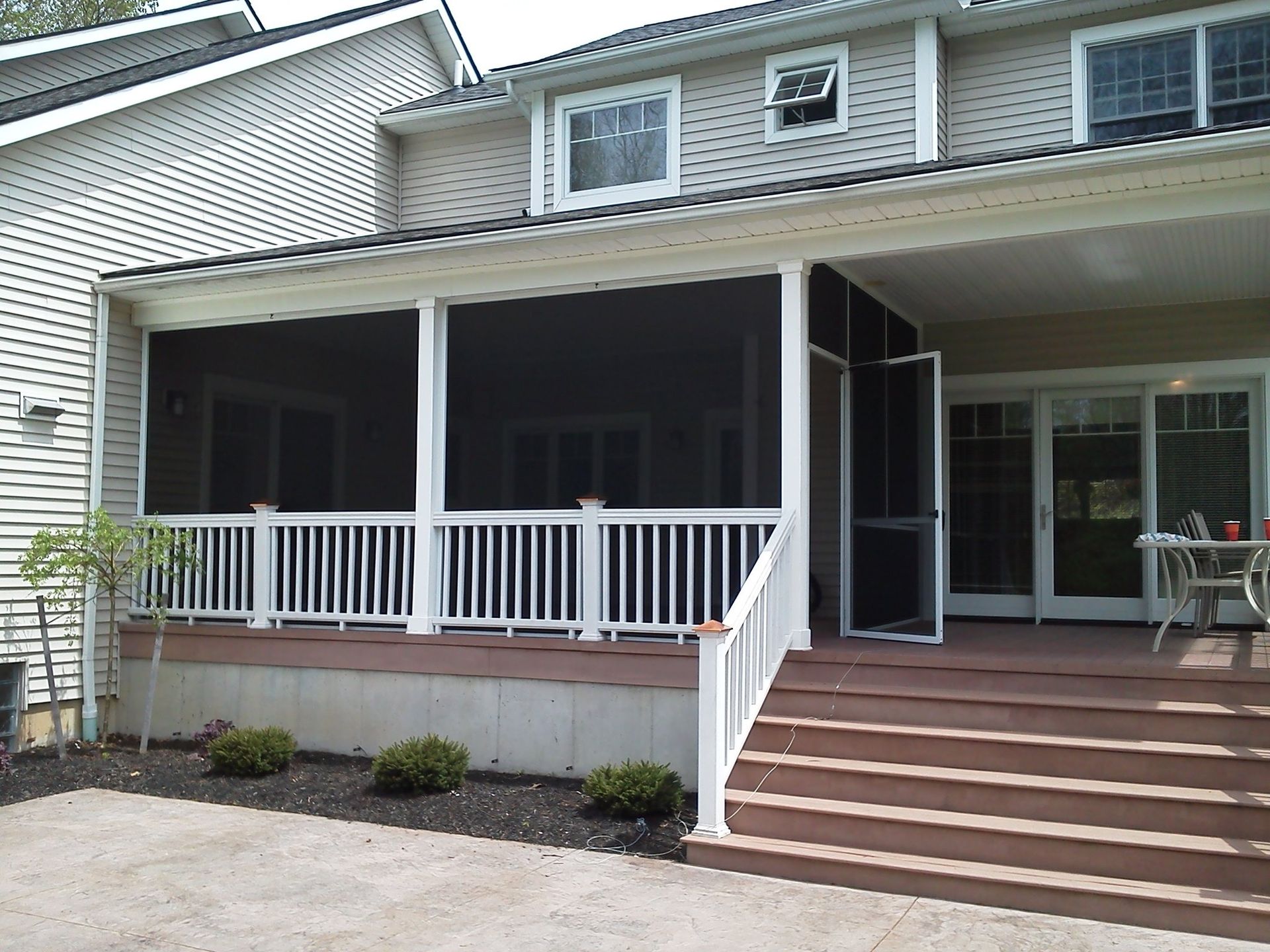 Screened porch with white railing, brown deck, and steps leading to a house with large windows.