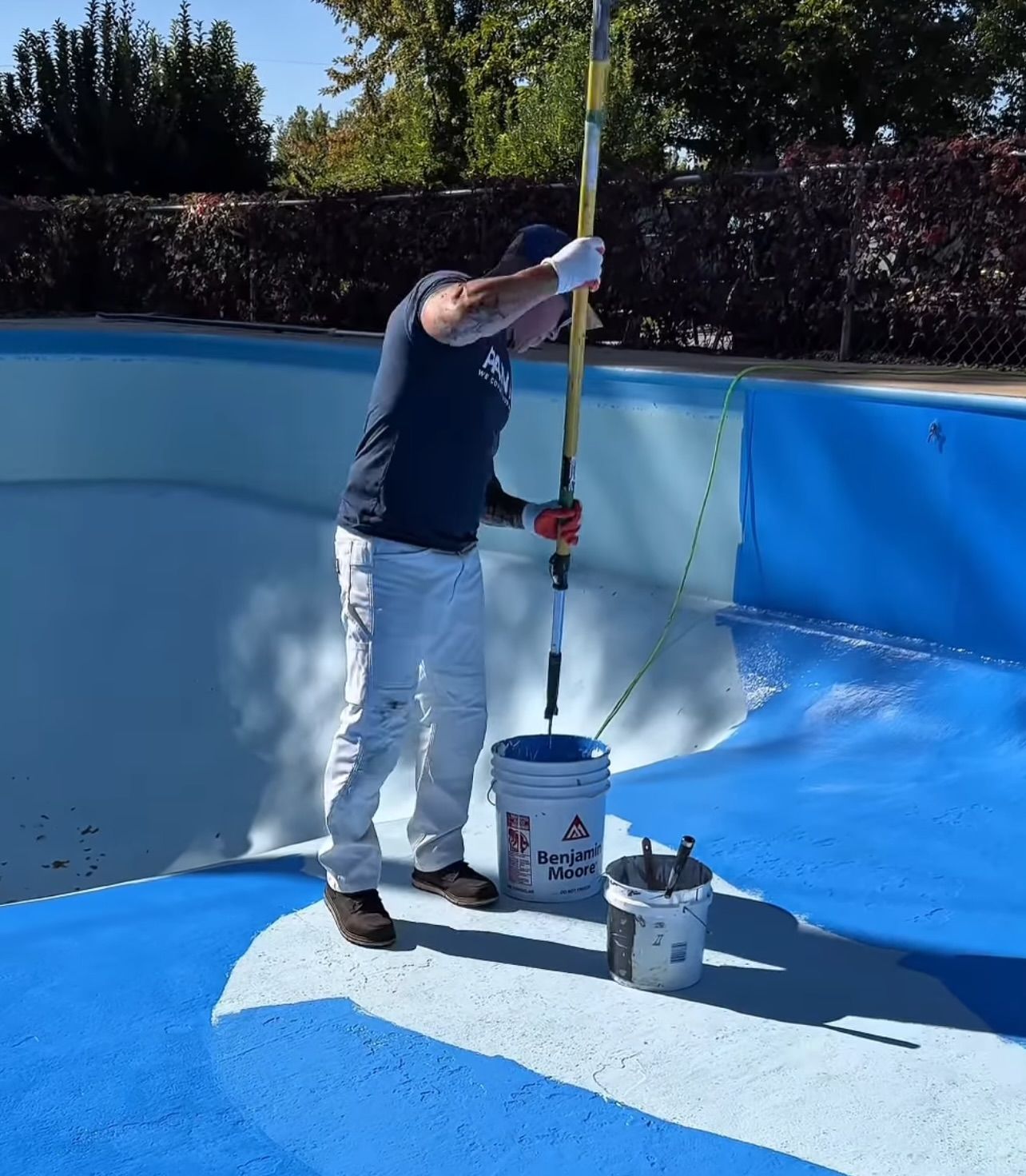 A man painting a blue swimming pool with a long-handled roller.