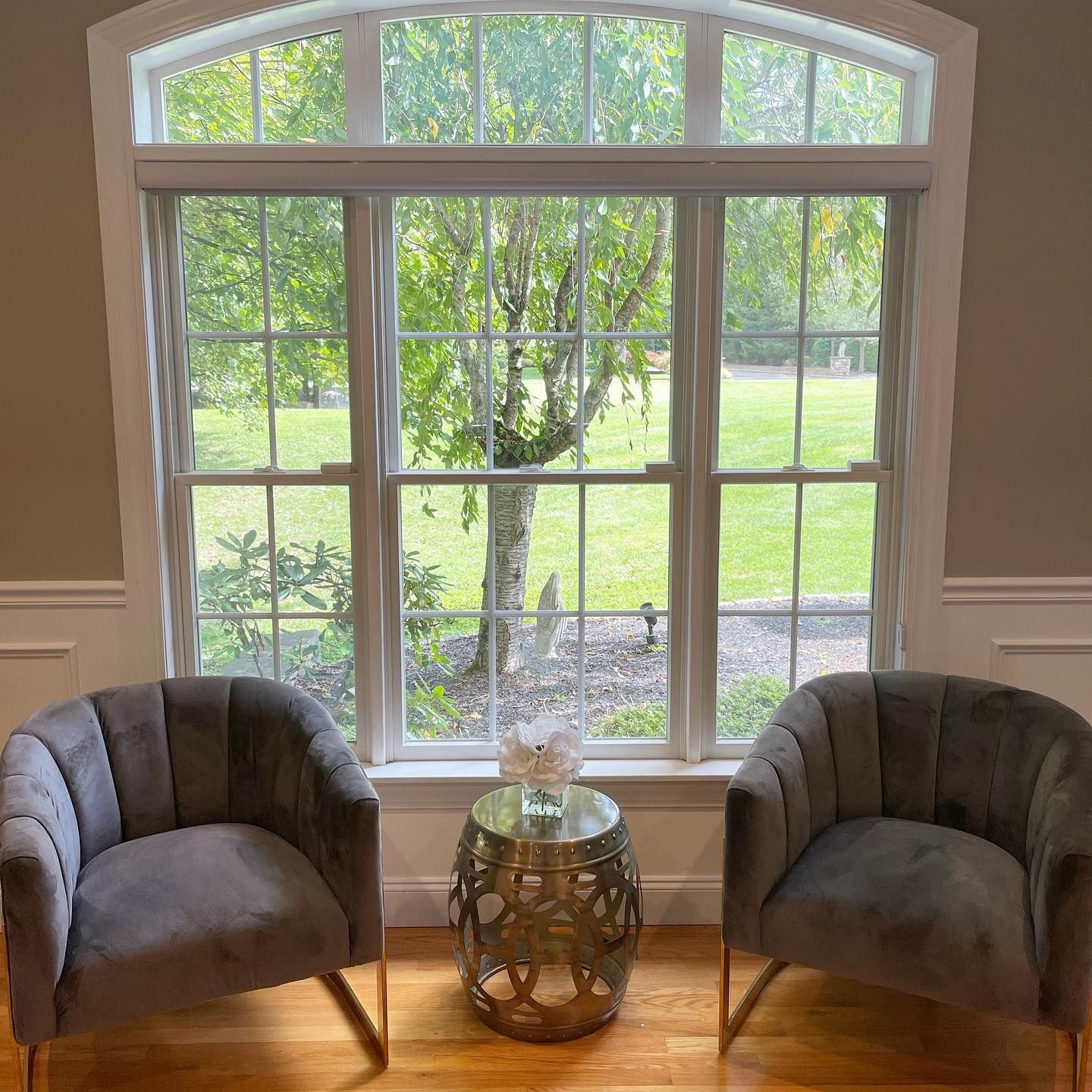 Two gray velvet chairs face a window with a small table in between. The chairs sit on a hardwood floor.
