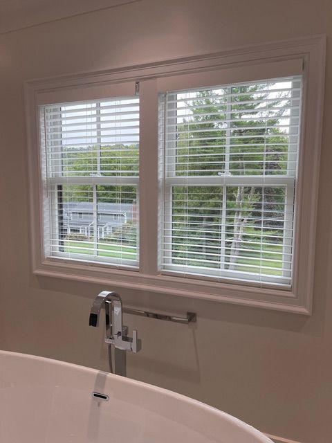 Bathroom window with white blinds, above a bathtub. Lush green trees are visible outside the window.