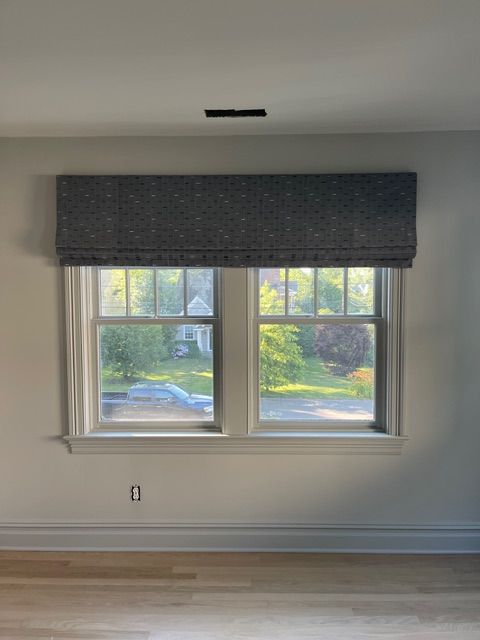 Window with two panes, gray Roman shade, view of trees and a house.