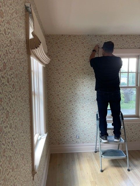 Man on a ladder installing wallpaper on a wall with a window. The room has floral wallpaper and wooden floors.