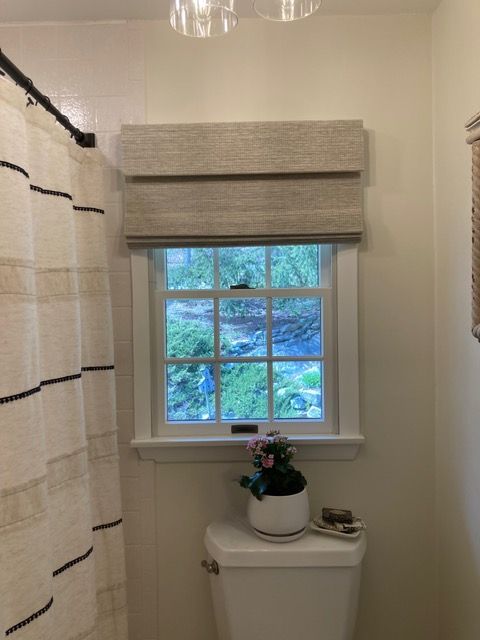 Bathroom with a window above a toilet; neutral colored roman shade; shower curtain to the left.