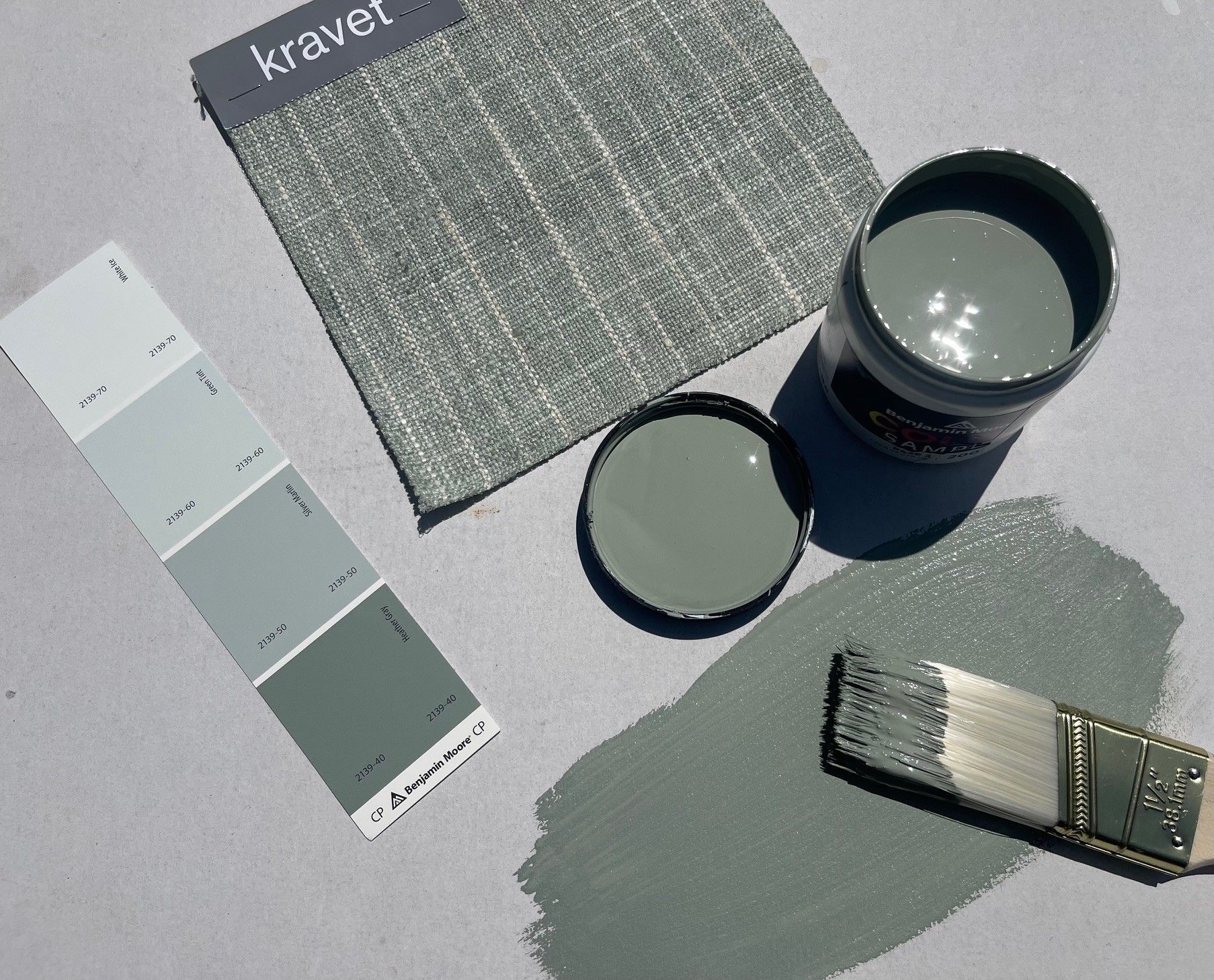 Fabric swatch, paint can, color palette, and paintbrush. Gray-green paint color samples on white surface.