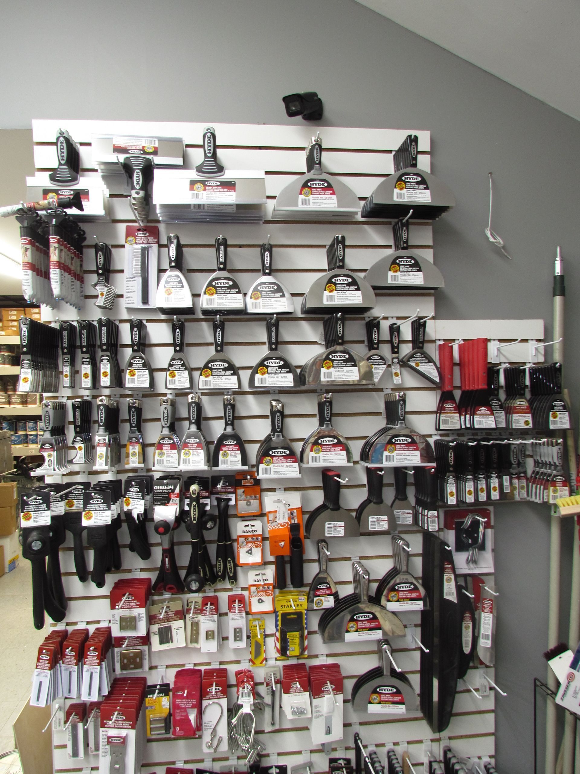 Tool display in a hardware store. Various putty knives, scrapers, and hand tools are organized on a white slatwall.