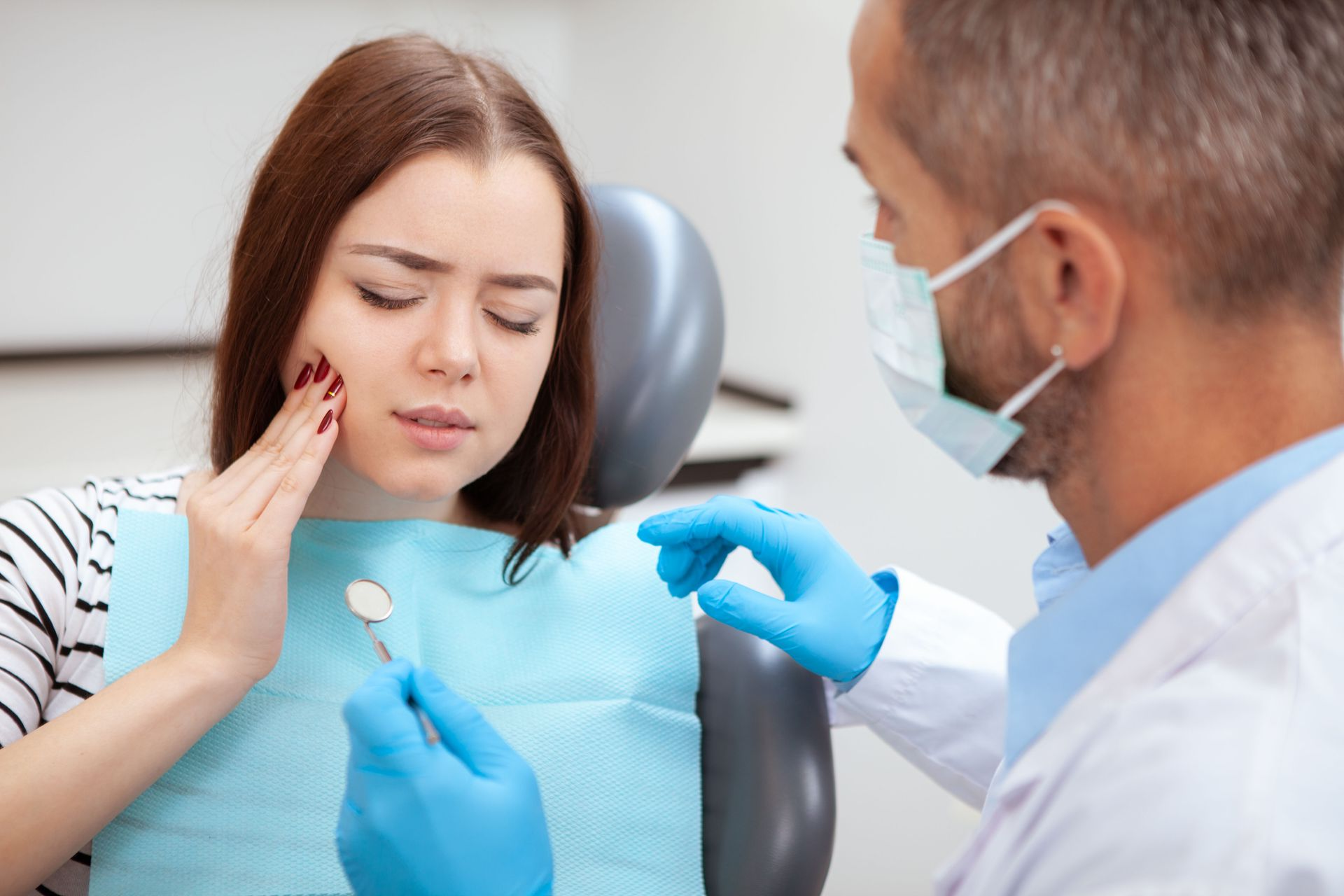 Woman with a pained expression at dentist, touching cheek. Dentist with mask examining, holding mirror.