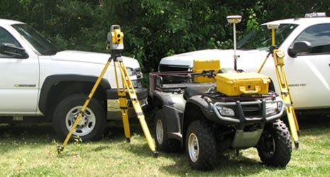 Surveying equipment, including a total station and GPS units on tripods, with an ATV and two pickup trucks in a field.