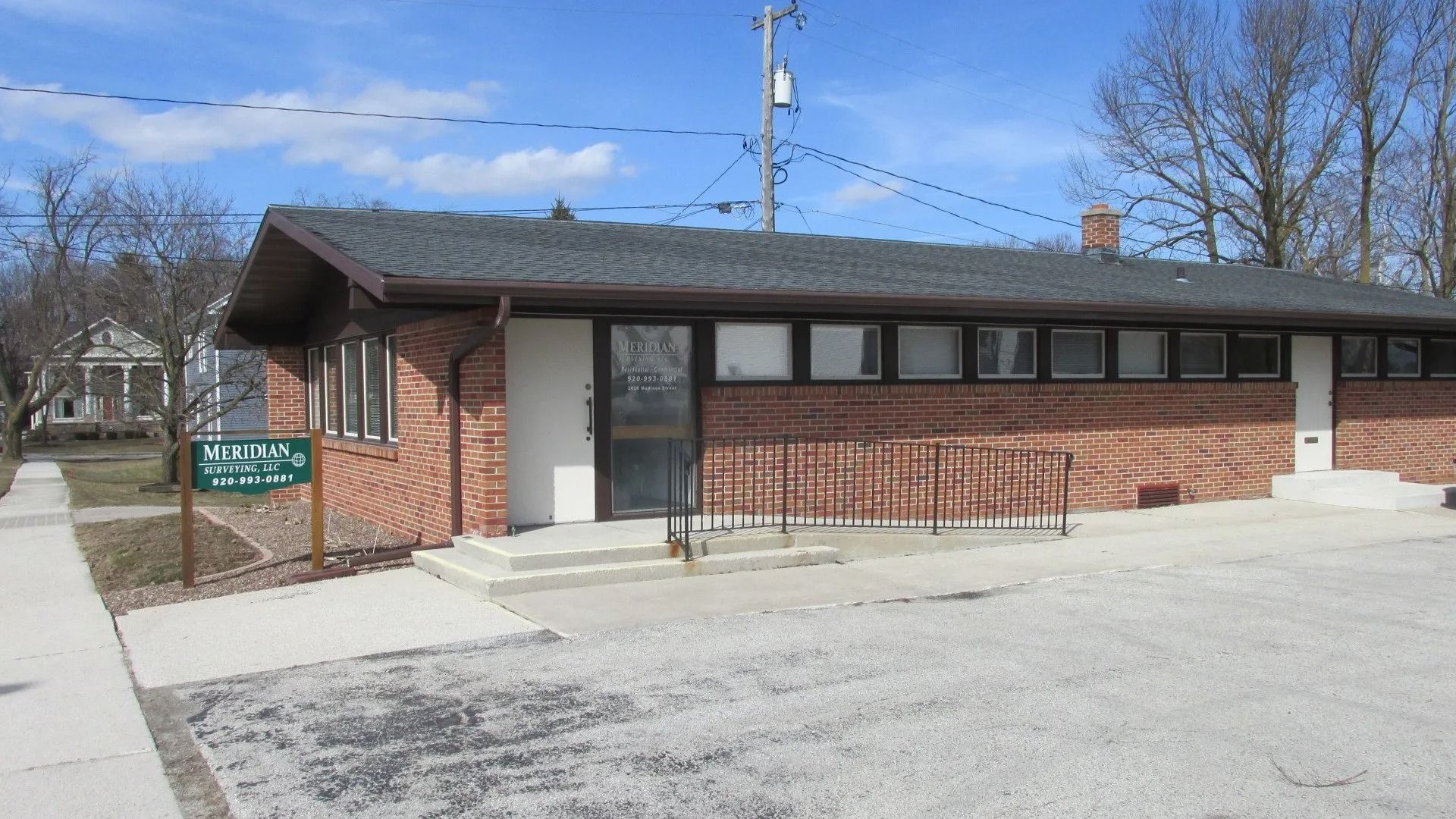 A single-story brick building with a dark shingled roof, a glass entrance, and a green sign in a paved lot.
