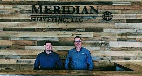 Two people stand behind a wooden counter in front of a rustic wood-plank wall featuring the Meridian Surveying, LLC logo.