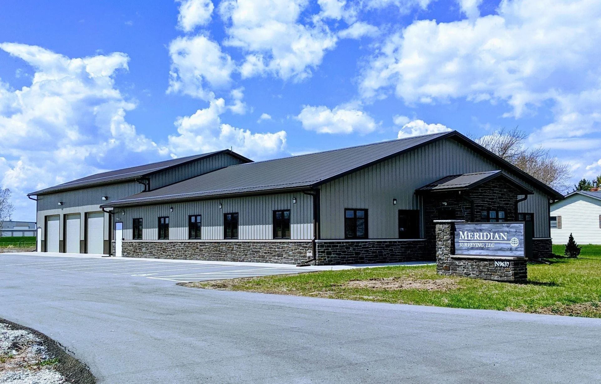 A tan metal commercial building with a stone foundation, garage doors on the side, and a sign in the front yard.
