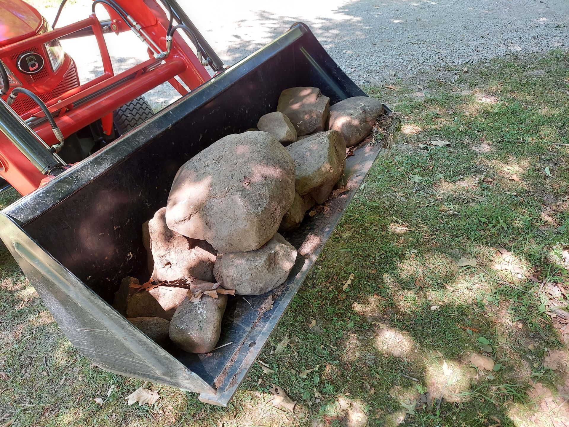 A bucket of rocks is sitting on the grass next to a tractor.