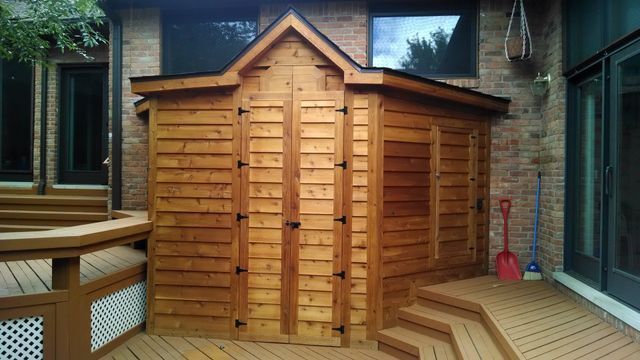 A wooden shed is sitting on a deck next to a brick building.