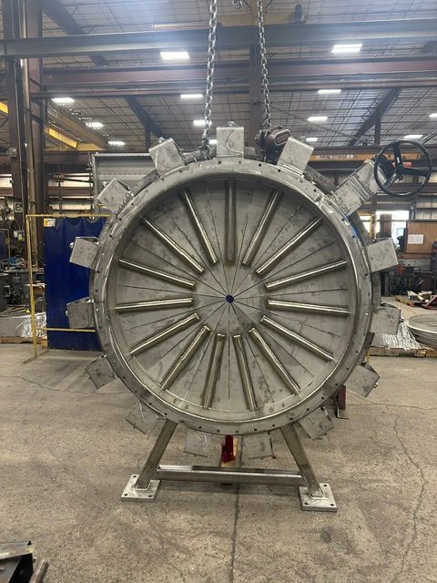 Large metal industrial component with radial bars and circular design, supported on a stand, suspended from overhead crane.