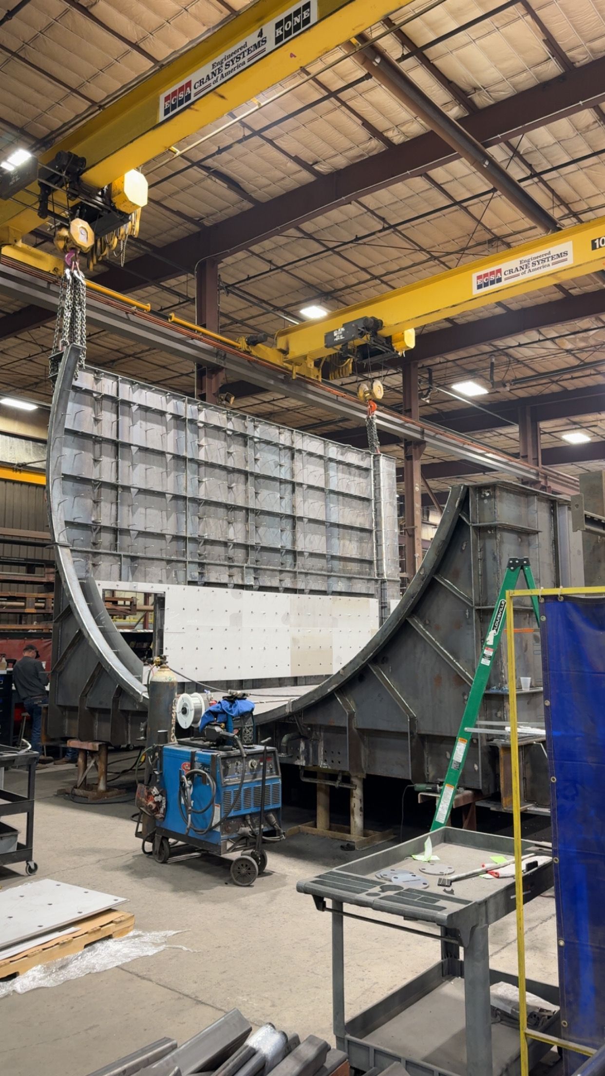 Large metal industrial structure being assembled in a workshop, under a yellow crane.